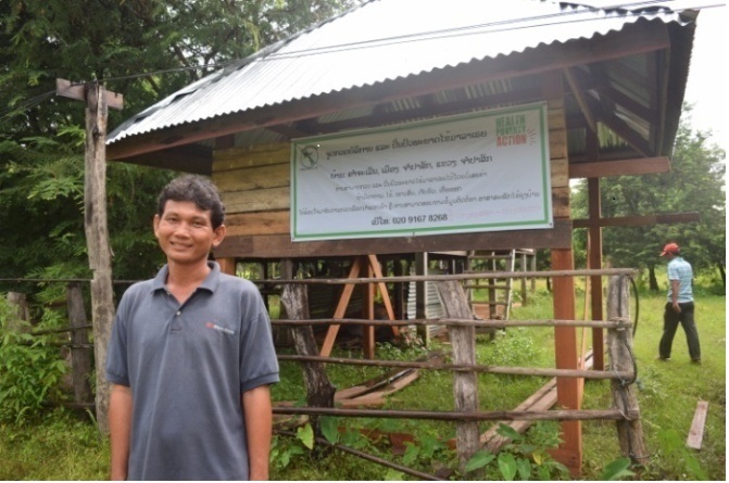 Douangpi stands in front of an awareness sign, which gives malaria information in a selection of local languages.