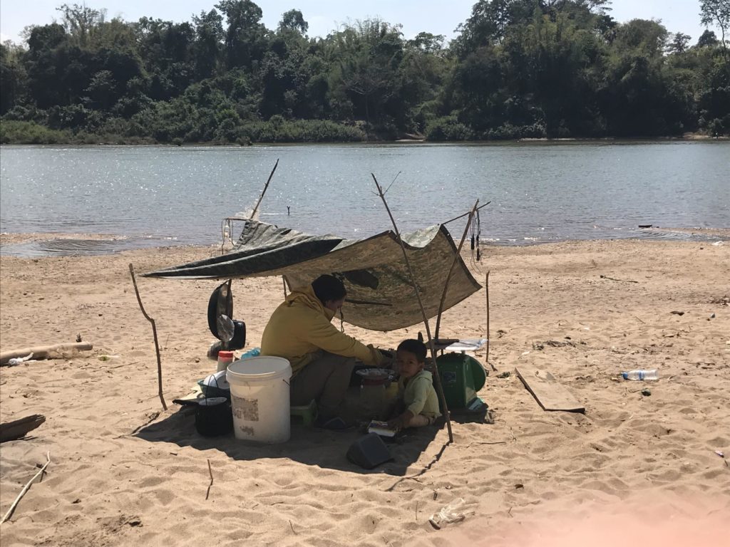 A man and child shelter from the sun under a tarpaulin on the sandy shore of a river